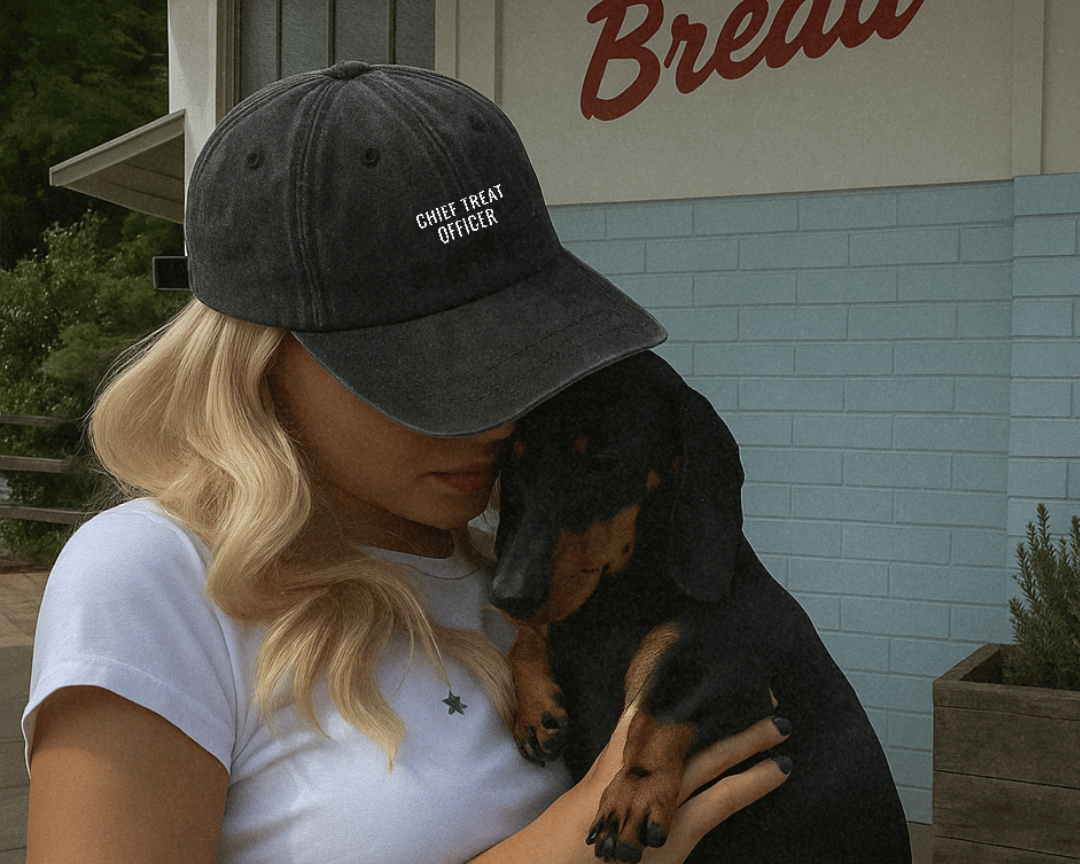 Person wearing a cap holding a small black and brown dog in front of a building. Her cap is embroidered 'chief treat officer'