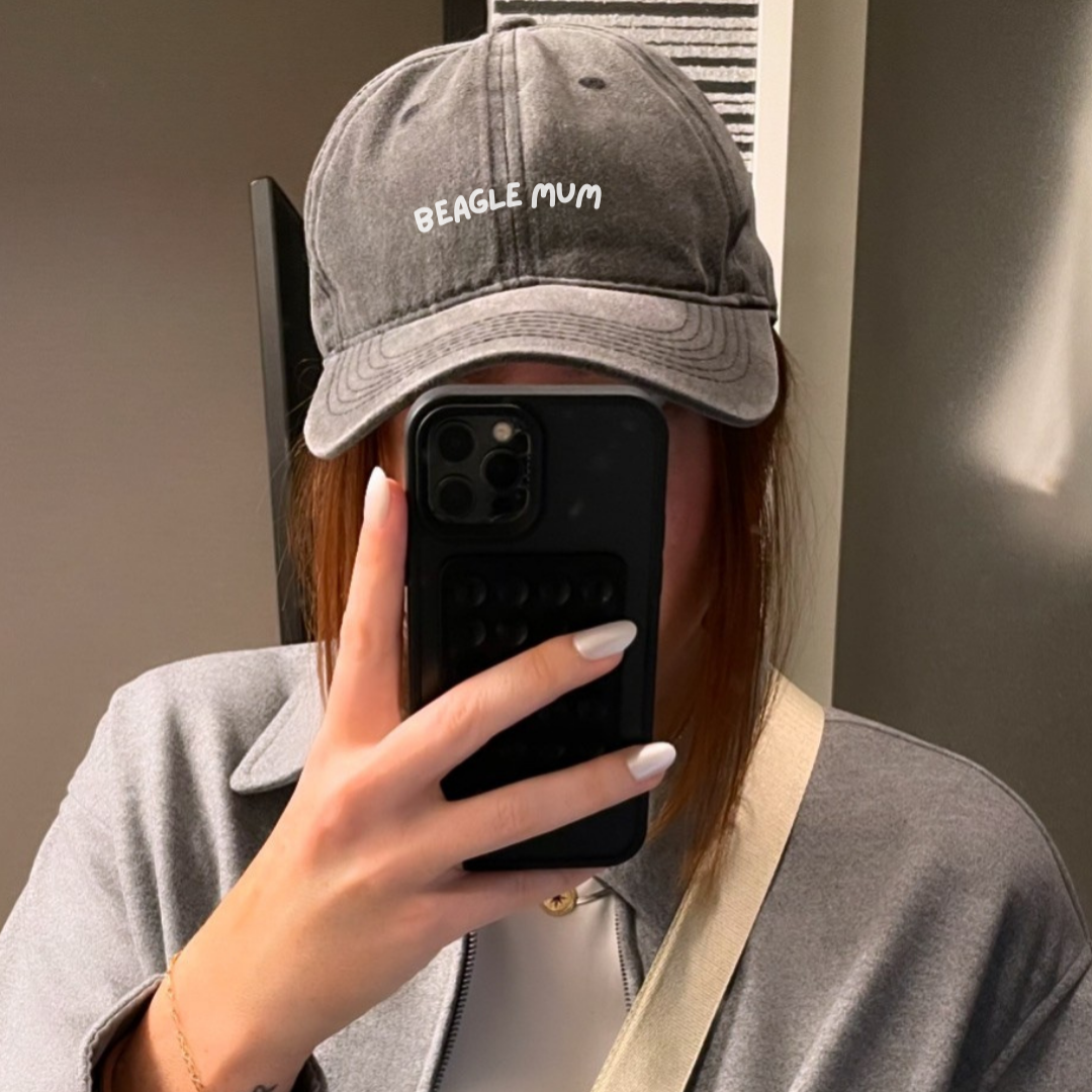 A person holding a smartphone in front of their face, wearing a gray baseball cap with 'BEAGLE MUM' text embroidered on the front.