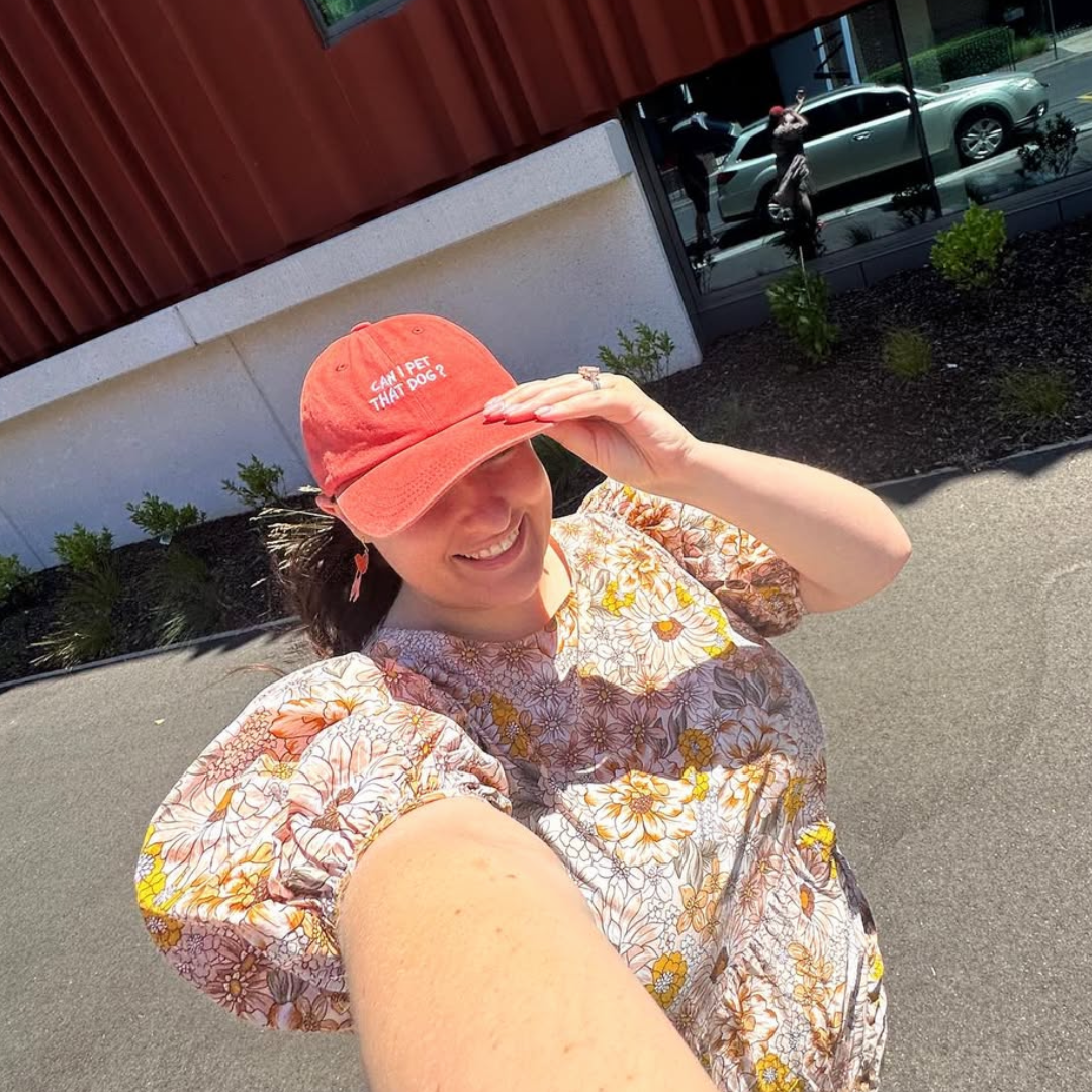 A person wearing a floral patterned shirt and an orange cap with text on it, standing outdoors.