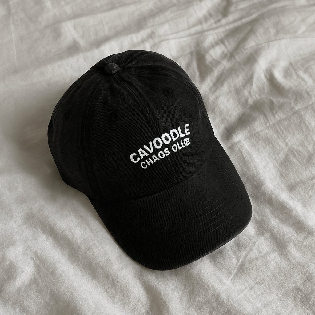 black denim cap with embroidered white text 'cavoodle chaos club' laying on a neutral linen cloth