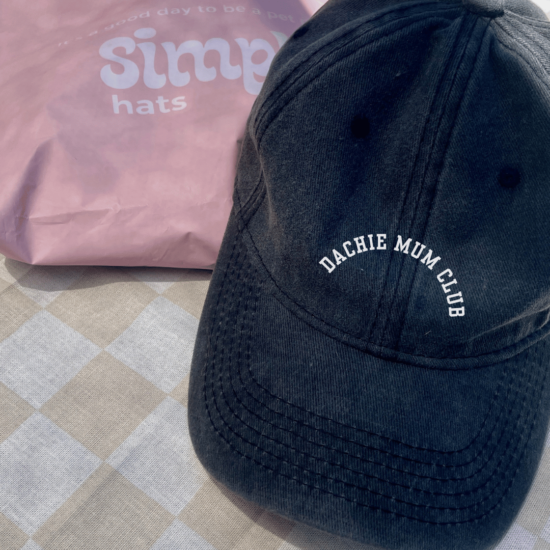 black vintage denim cap with white curved embroidered writing "Dachie Mum Club" it is on a checkered tablecloth with a pink posting satchel from Simply Hats