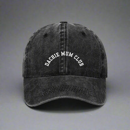 A black baseball cap with 'Dachie Mum Club' embroidered on the front, placed on a checkered surface, with a pink item in the background.