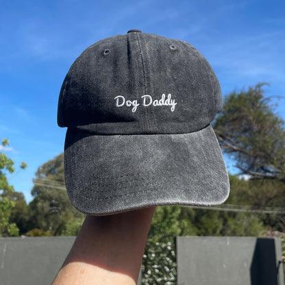 A gray baseball cap with 'Dog Daddy' embroidered on the front, held up against a sky background.
