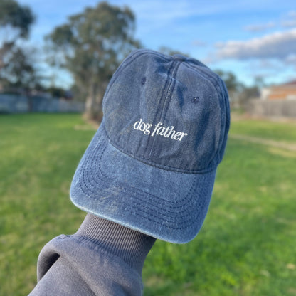 A blue denim baseball cap with 'dog father' text embroidered on the front, held up against an outdoor background.