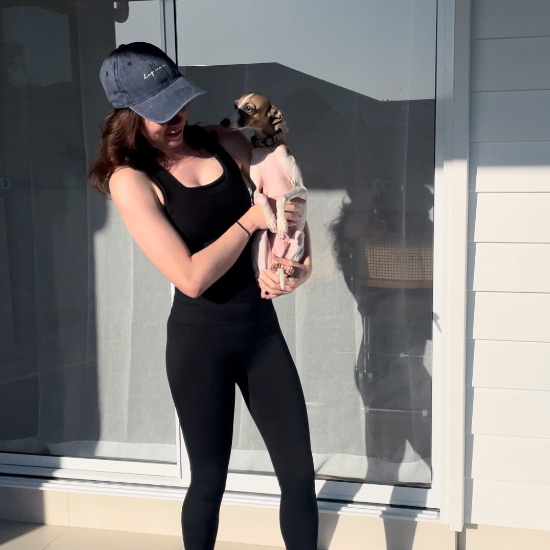 Woman in black athletic wear holding a small dog outdoors. Her cap is black and embroidered "Dog mum"