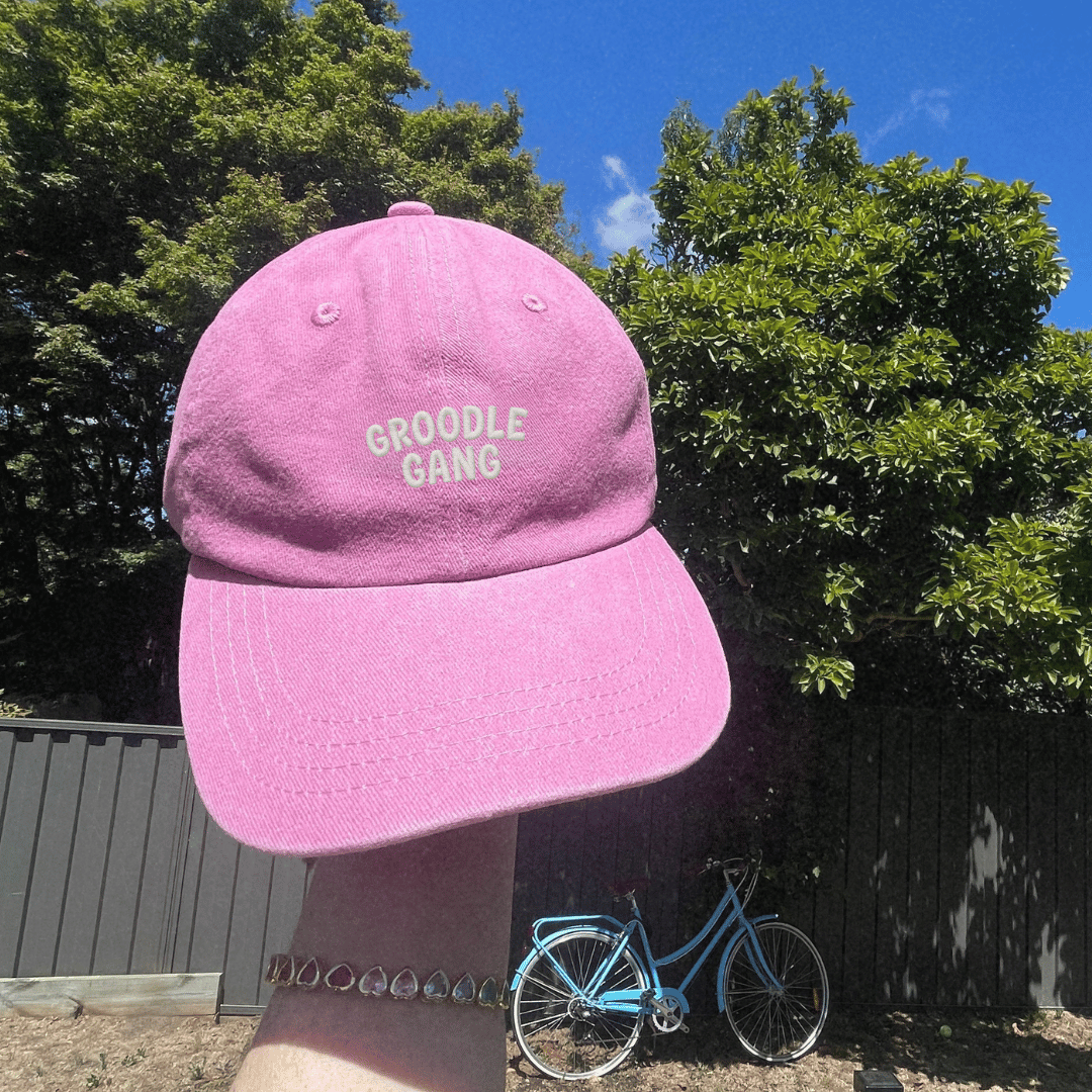 Pink cap with 'GROODLE GANG' text against a natural background with trees and a bicycle.