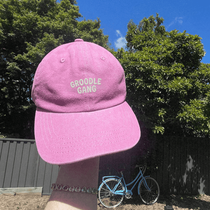 Pink cap with 'GROODLE GANG' text against a natural background with trees and a bicycle.