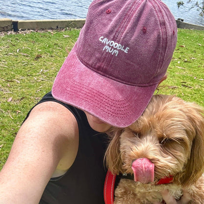 A person wearing a pink 'Cavoodle Mum' cap with an adjustable metal clasp, sitting beside a brown and white dog with a red collar.