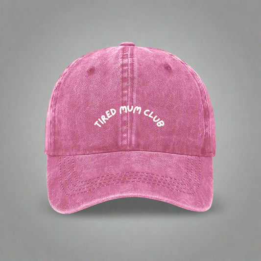 A pink baseball cap with 'TIRED MUM CLUB' embroidered in white on the front.