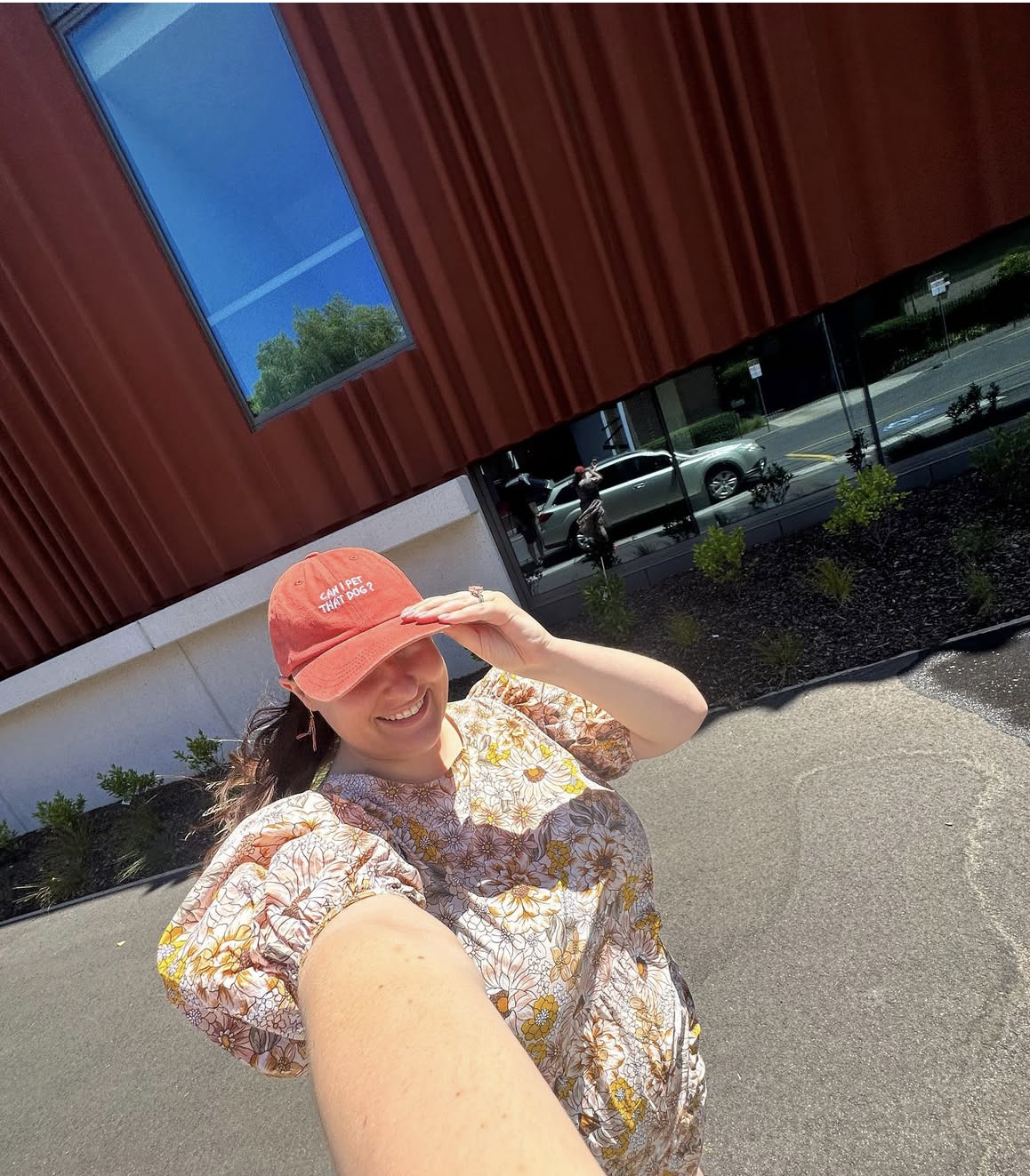 Person wearing a red cap taking a selfie outdoors with a building and street in the background. Her cap is embroidered "can i pet that dog?"