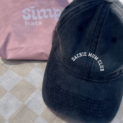 A black baseball cap with 'Dachie Mum Club' embroidered on the front, placed on a checkered surface, with a pink item in the background.