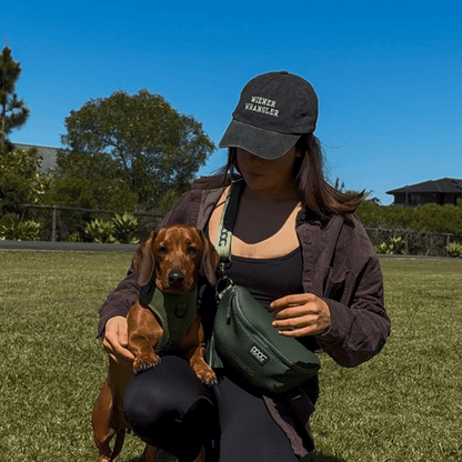 Person holding a small dog in a park with a clear blue sky. her cap says Wiener Wrangler