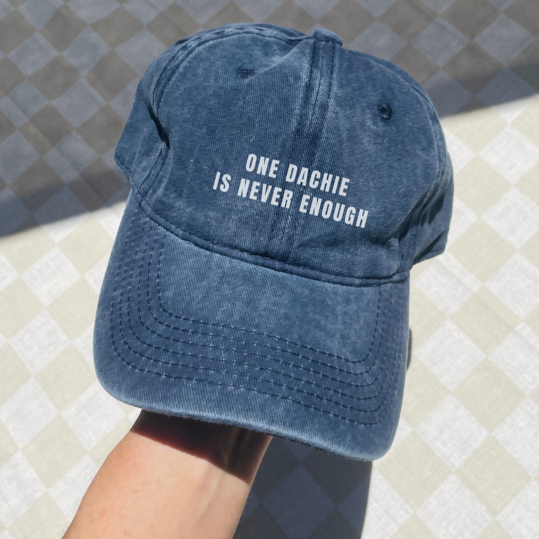 A blue denim baseball cap with the text 'ONE DACHIE IS NEVER ENOUGH' printed in white on the front.