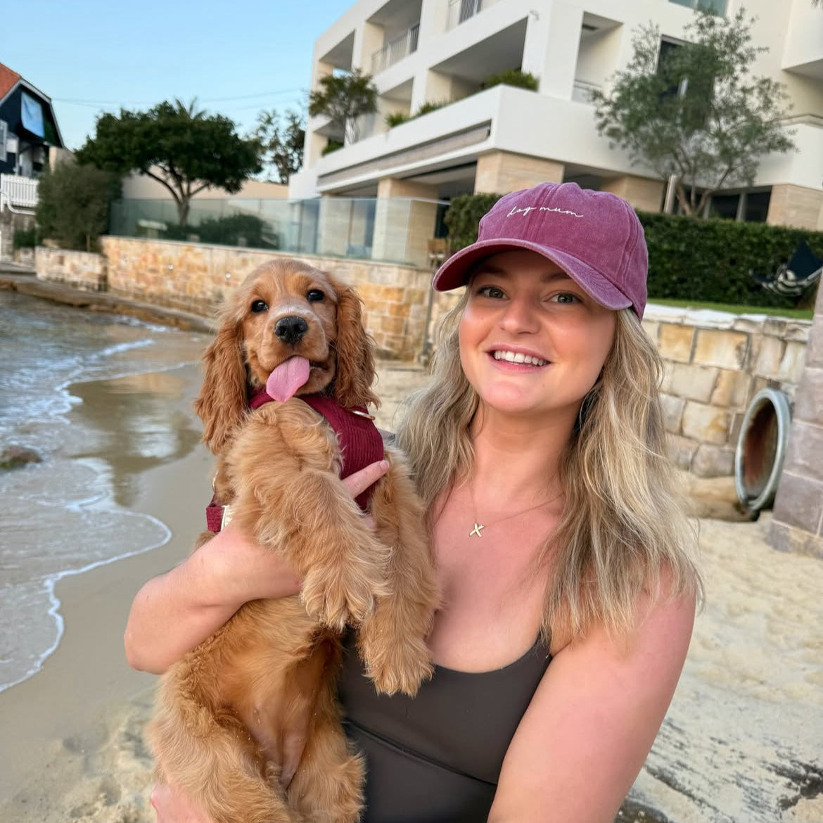 Woman holding a dog on a beach with buildings in the background. her cap is maroon and is embroidered "dog mum"