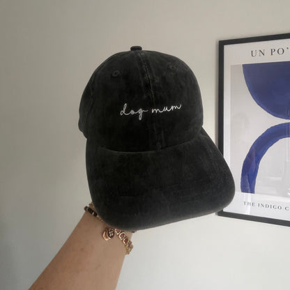 A black baseball cap with the text 'dog mum' embroidered on the front, held up against a wall.