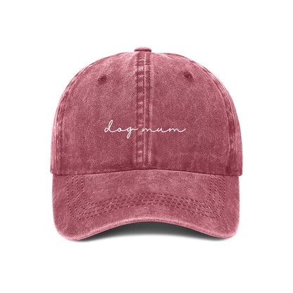 A maroon baseball cap with the text 'dog mum' embroidered on the front, held up against a wall.