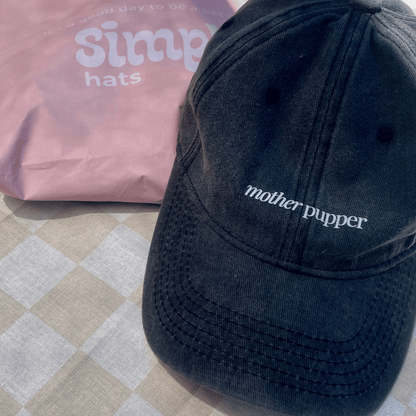 A black denim cap with the text 'mother pupper' embroidered on the front, placed on a patterned surface, with a pink object in the background.