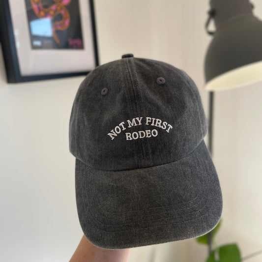 Black cap with 'Not My First Rodeo' text held by a hand against a neutral background