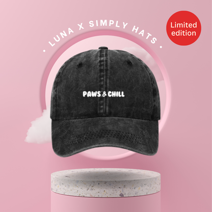 A gray baseball cap with 'PAWS & CHILL' written in white text on the front, held in a person's hand.
