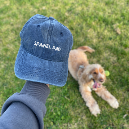 A denim baseball cap with the text 'SPANIEL DAD' on it, held in a person's hand with a brown dog lying on grass in the background.
