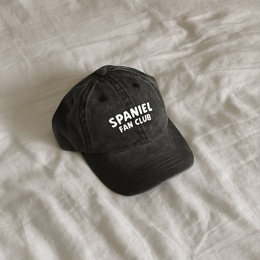 Black cap with 'SPANIEL FAN CLUB' text on a beige fabric background