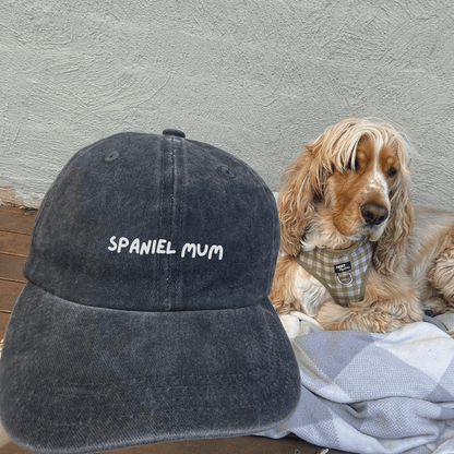 A denim cotton cap with 'SPANIEL MUM' text embroidered on the front, lying next to a cocker spaniel dog with a bandana.