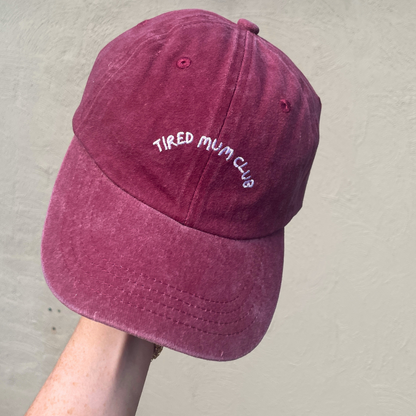A maroon baseball cap with 'TIRED MUM CLUB' embroidered in white on the front.