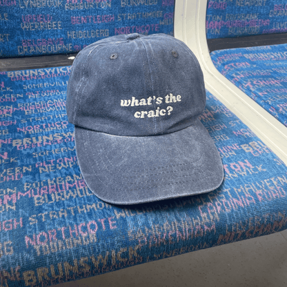 A denim baseball cap with the text 'what's the craic?' printed on the front, placed on a surface with text patterns.