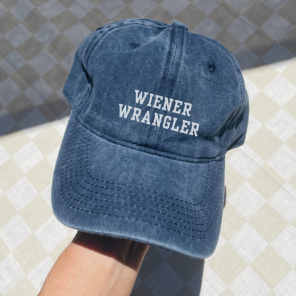 A blue denim baseball cap with 'WIENER WRANGLER' text embroidered on the front.